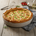 A baked salmon and green pepper quiche in a round ceramic dish, set on a rustic wooden table with a gray cloth, a whisk, and fresh herbs.
